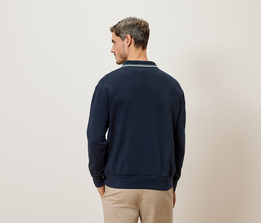 Homme vu de dos, portant un sweat-shirt marine à manches longues avec un col vert clair et un pantalon beige.