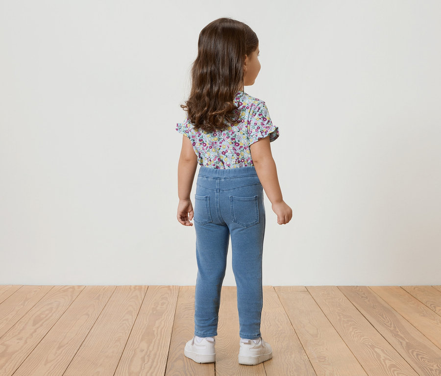 Vue arrière d'une petite fille aux cheveux bouclés portant un haut bleu à fleurs à manches courtes, un legging bleu et des baskets blanches.