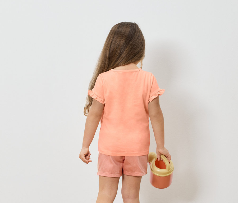 Fillette vue de dos portant un t-shirt et un short roses, tenant un arrosoir orange.