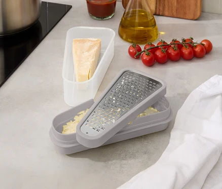 Sur un comptoir de cuisine, il y a une casserole, de la sauce tomate, une bouteille d'huile, des tomates, une râpe à fromage avec du fromage et un morceau de fromage dans une boîte.