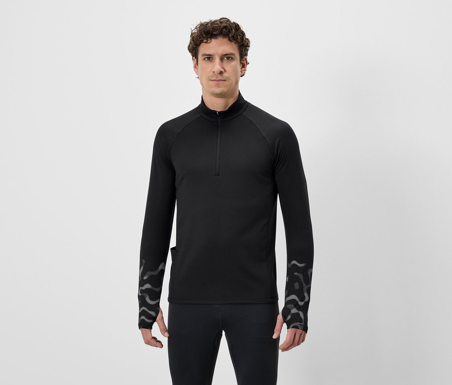Un homme aux cheveux bouclés porte un t-shirt noir à manches longues avec fermeture éclair et un legging sur fond blanc.