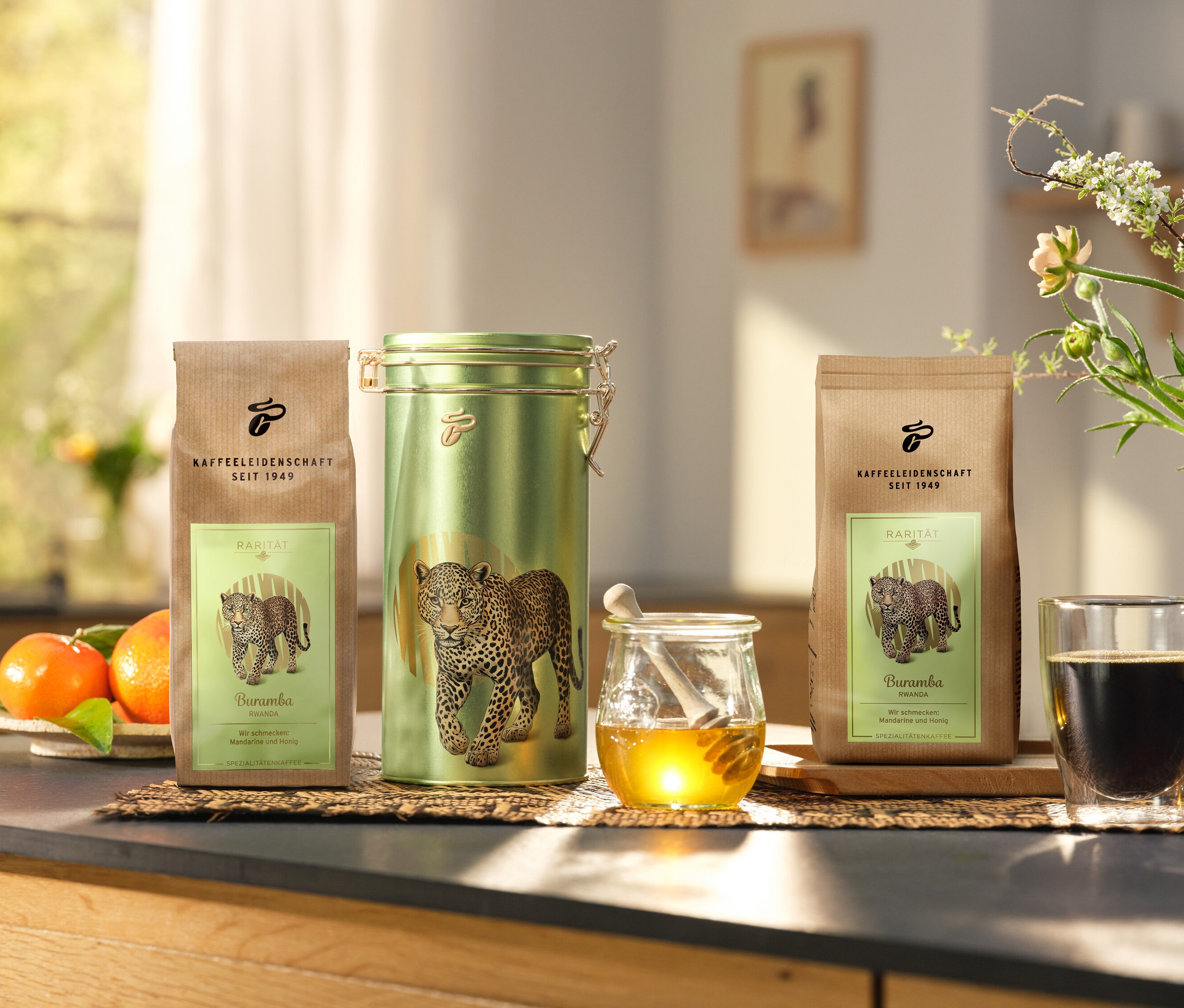 Sur un comptoir de cuisine, une boîte à café et deux paquets de »Buramba Rwanda« avec boîte à café sont posés à côté d'une tasse de café et de miel.