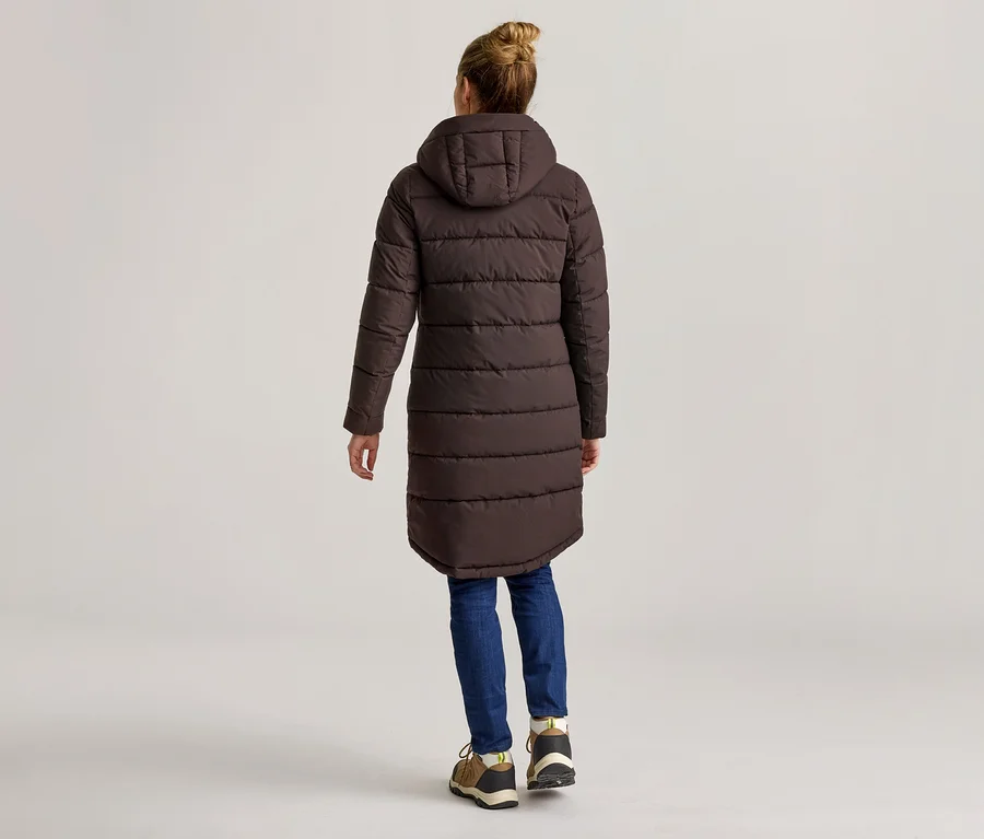 Femme vue de dos portant un long manteau matelassé marron à capuche, un jean bleu et des chaussures marron sur fond clair.