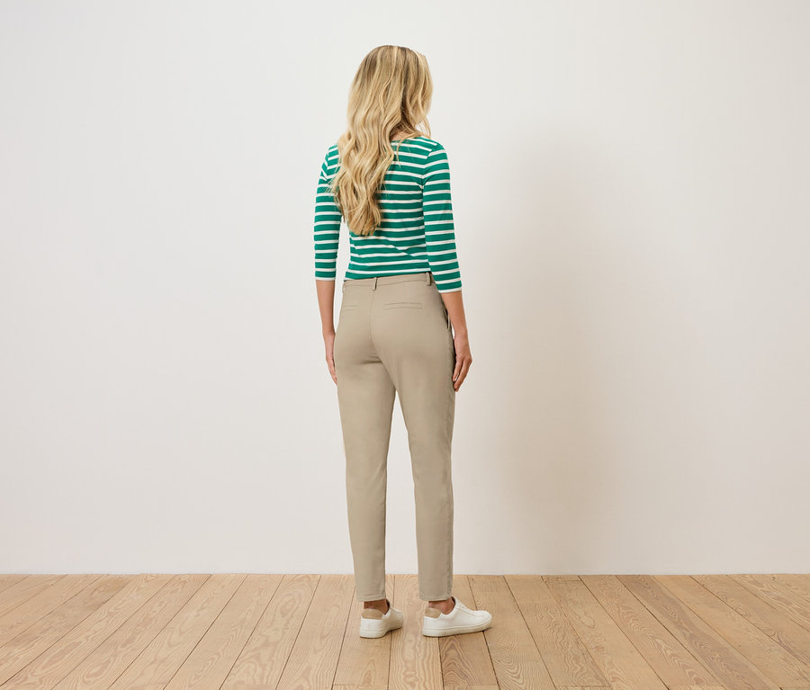 Une femme aux cheveux blonds et ondulés se tient dos au spectateur, portant un haut rayé vert et blanc, un pantalon beige et des baskets blanches.