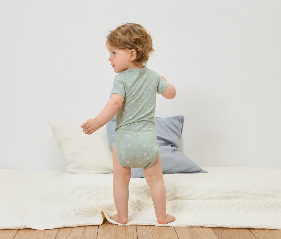 Un bébé en body vert clair à motifs de cœurs se tient debout sur un tapis d'éveil blanc.