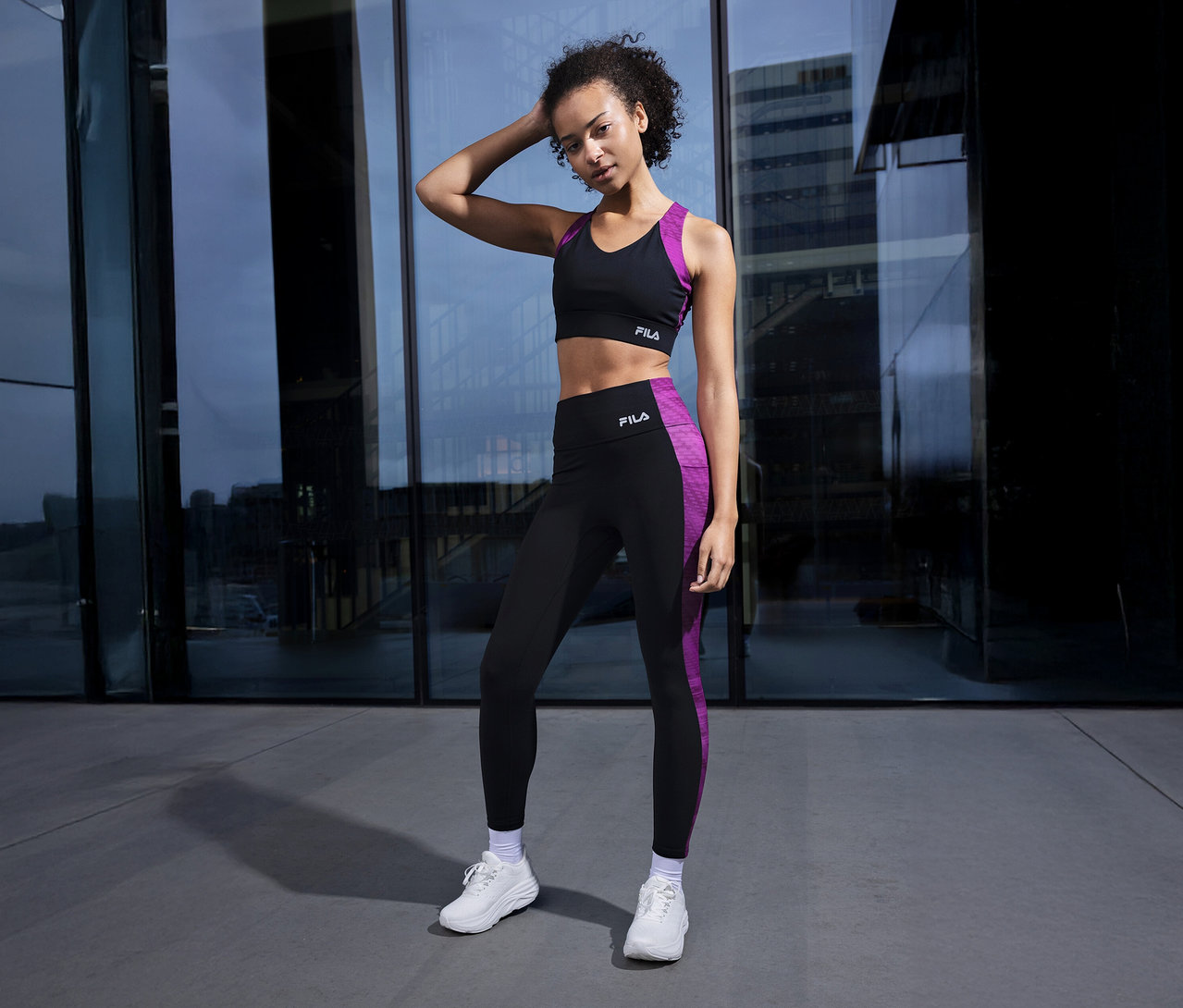 Femme posant devant une façade en verre, portant une soutien-gorge de sport FILA noir avec des détails violets et des leggings assortis.