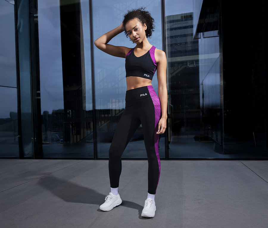 Femme posant devant une façade en verre, portant une soutien-gorge de sport FILA noir avec des détails violets et des leggings assortis.