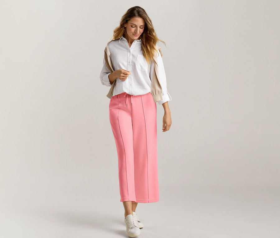 Une femme aux longs cheveux bruns avance en regardant vers le bas. Elle porte une chemise blanche, un pull beige drapé sur ses épaules et un pantalon en mélange de modal rose corail, associé à des baskets blanches.