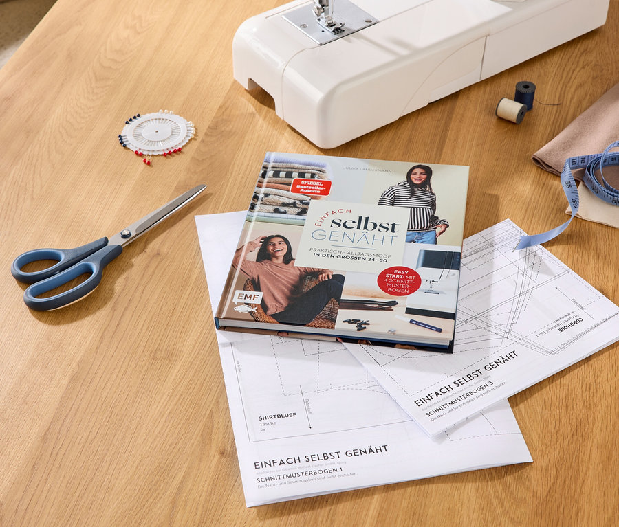 Machine à coudre, livre « Einfach selbst genäht », patrons de couture, ciseaux, coussin à épingles, fil, mètre ruban et tissu sur une table en bois.