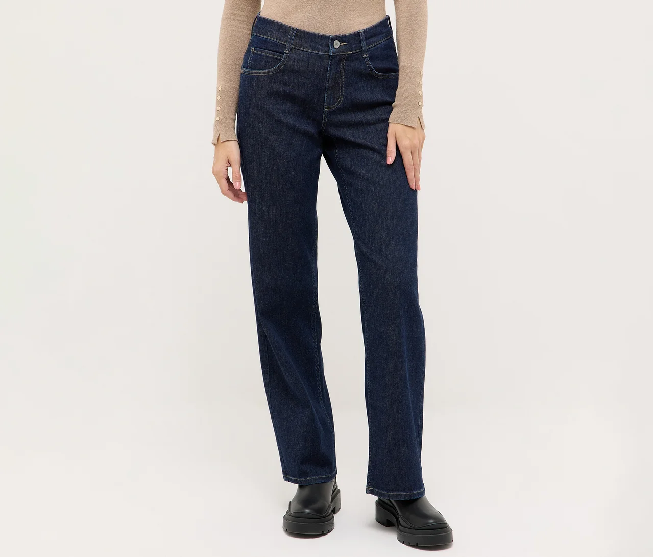 Personne portant un jean droit bleu foncé, un haut beige à manches longues avec des détails boutonnés aux poignets et des bottes noires à semelle épaisse.