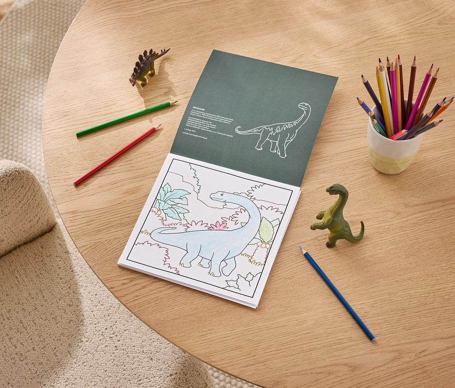 Un livre de coloriage ouvert sur une table en bois montre un dinosaure bleu partiellement colorié. À côté se trouvent un pot de crayons de couleur, des crayons rouges, verts et bleus, et deux jouets dinosaures.