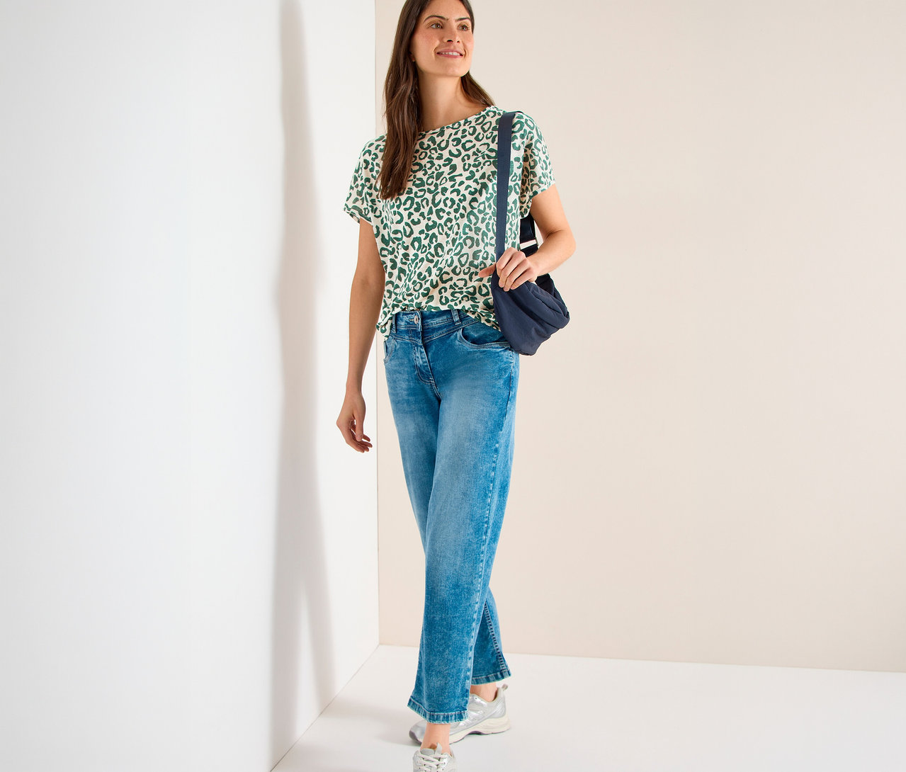 Une femme souriante porte un T-shirt manches courtes CECIL beige à motif léopard vert, un jean bleu, des baskets argentées et un sac bandoulière bleu foncé.