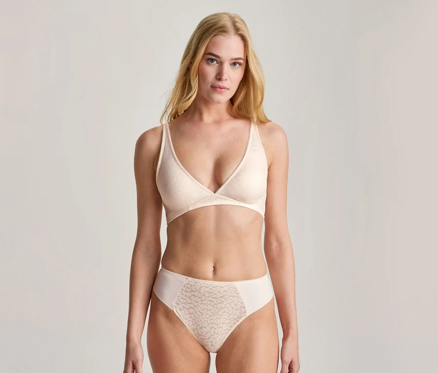 Une jeune femme aux longs cheveux blonds, vêtue d'un soutien-gorge souple beige clair et d'une culotte assortie avec un motif en dentelle subtil, regarde directement l'appareil photo.