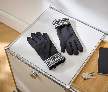 Une paire de gants noirs avec des poignets à motif pied-de-poule est posée sur une surface blanche. À côté se trouvent un smartphone et des clés sur un plateau en bois.