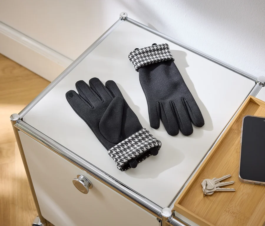 Une paire de gants noirs avec des poignets à motif pied-de-poule est posée sur une surface blanche. À côté se trouvent un smartphone et des clés sur un plateau en bois.