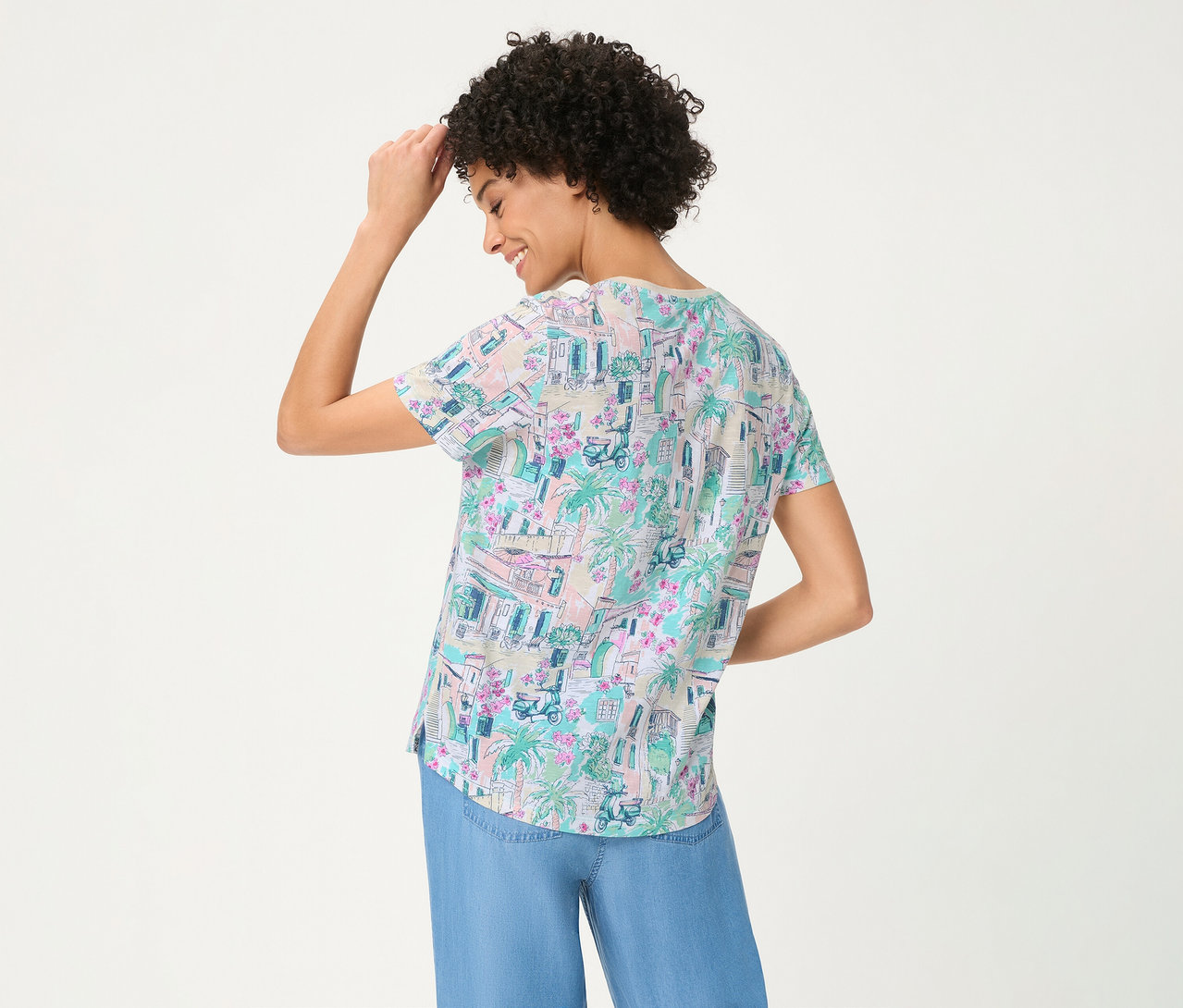 Femme de dos, souriante et touchant sa tête, portant un T-shirt Olsen à motif coloré de bâtiments, palmiers et scooters, associé à un pantalon large bleu.