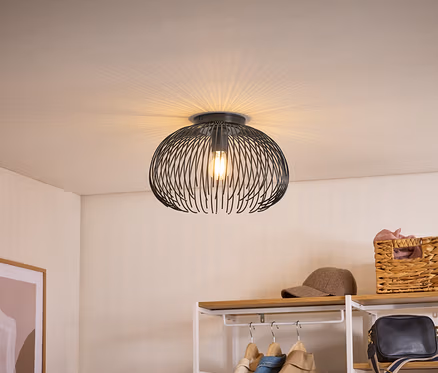 Plafonnier noir avec abat-jour en fil de fer, projetant une lumière en éventail sur le plafond. En dessous, une étagère avec vêtements, casquette et panier.
