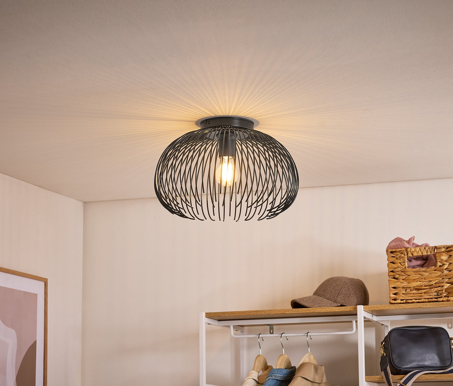 Plafonnier noir avec abat-jour en fil de fer, projetant une lumière en éventail sur le plafond. En dessous, une étagère avec vêtements, casquette et panier.