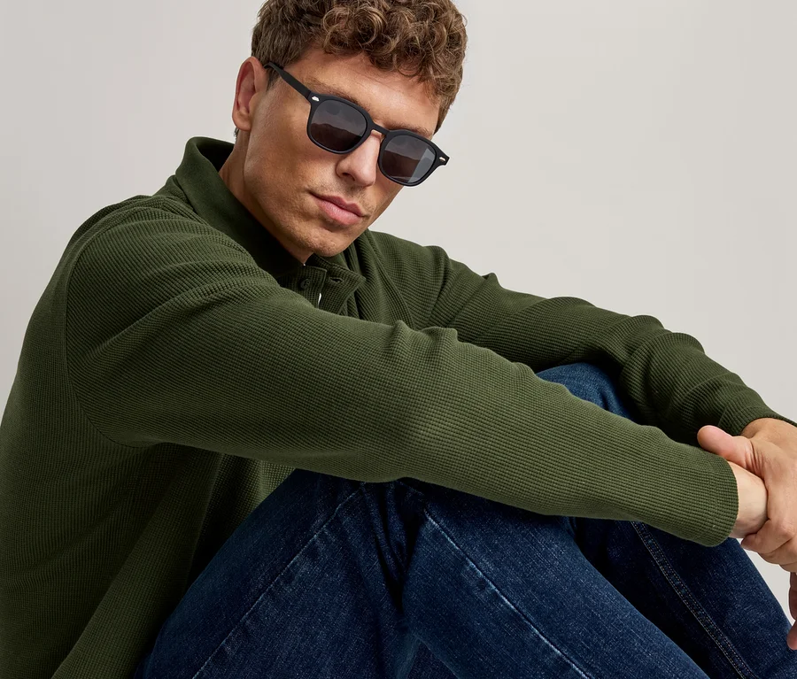 Homme portant des lunettes de soleil noires, un polo vert à manches longues et un jean bleu, assis et regardant devant lui.