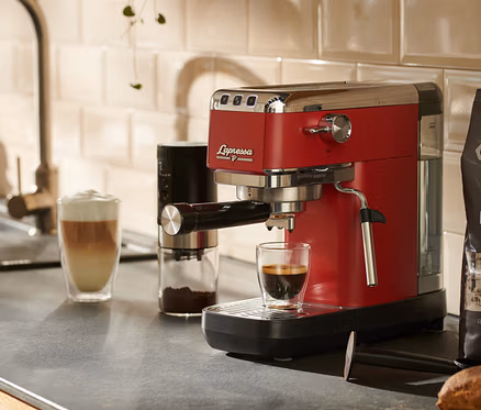 Une machine à porte-filtre « Lapressa » rouge est posée sur un plan de travail de cuisine à côté d'un verre de café.