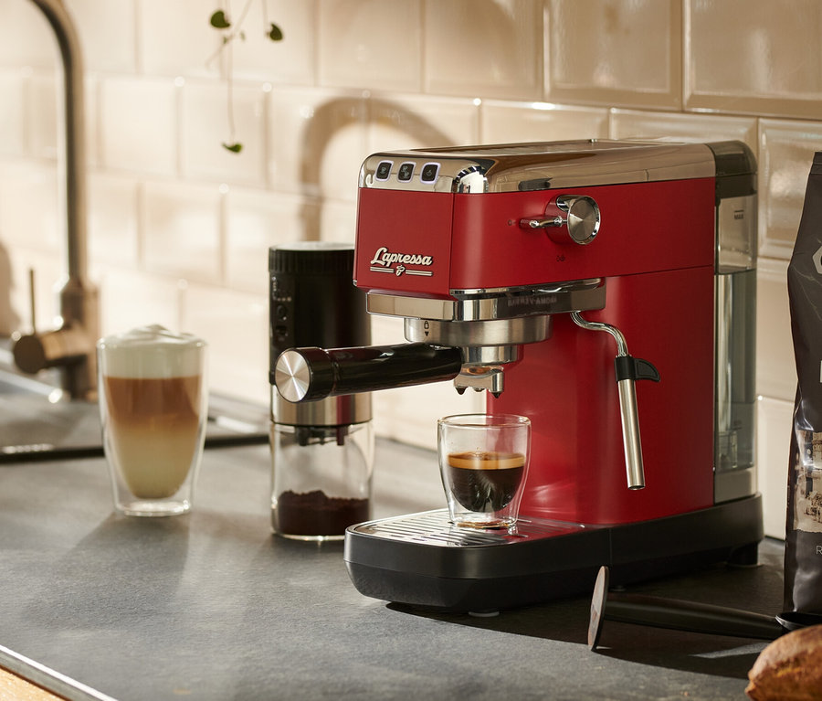 Une machine à porte-filtre « Lapressa » rouge est posée sur un plan de travail de cuisine à côté d'un verre de café.