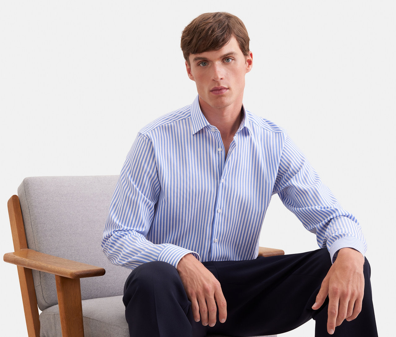 Homme assis dans un fauteuil gris, portant une chemise à manches longues rayée bleu et blanc et un pantalon foncé.