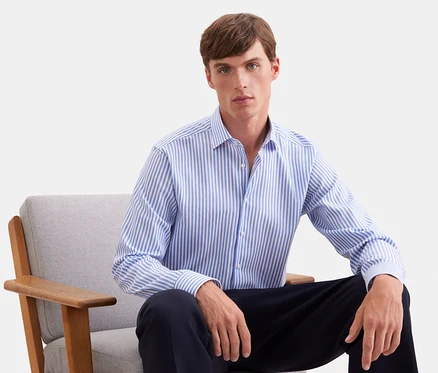 Homme assis dans un fauteuil gris, portant une chemise à manches longues rayée bleu et blanc et un pantalon foncé.