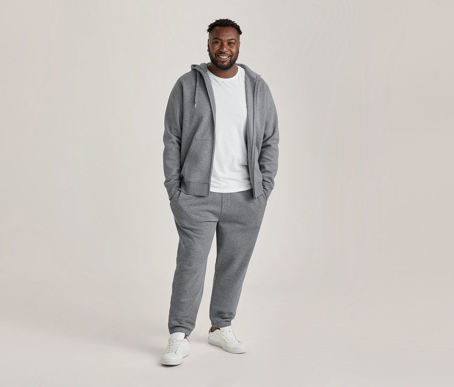 Homme souriant portant un sweat à capuche zippé gris, un t-shirt blanc à encolure ronde, un pantalon de jogging gris et des baskets blanches, les mains dans les poches.