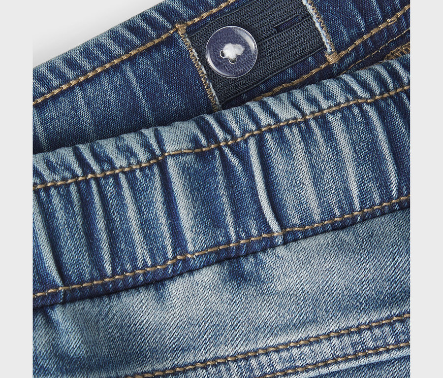 Gros plan d'un jean slim sweat NAME IT bleu, montrant la taille élastiquée avec un bouton transparent pour l'ajustement et des surpiqûres marron.