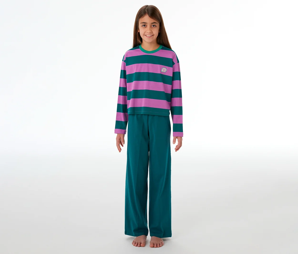 Une jeune fille souriante, pieds nus, porte un pyjama rayé violet et turquoise avec un pantalon assorti.