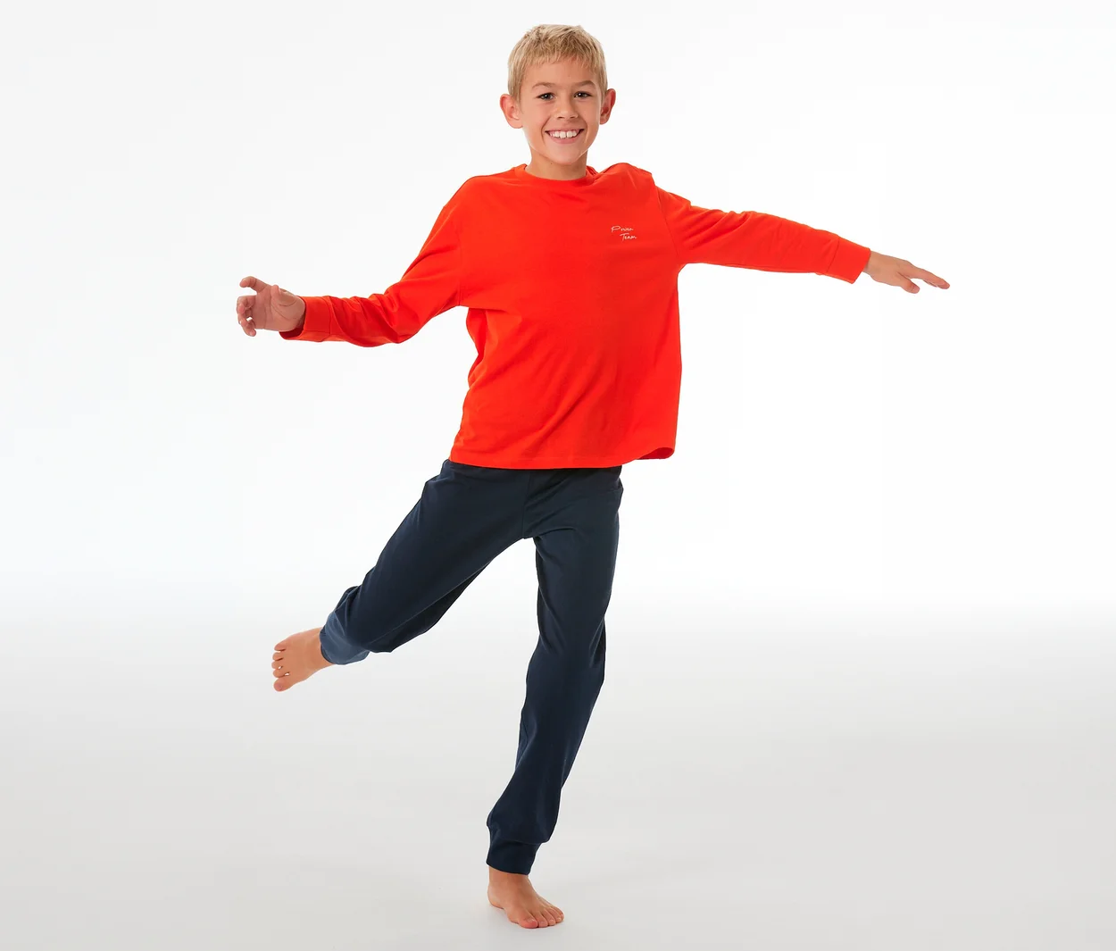 Garçon souriant en équilibre sur une jambe, portant un haut de pyjama orange à manches longues et un pantalon de pyjama enfant bleu foncé avec des poignets.