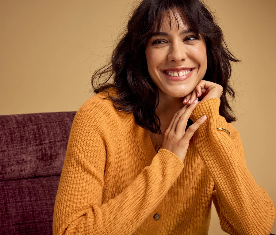 Femme souriante aux cheveux foncés portant un cardigan en maille orange et un bracelet doré, les mains jointes sous le menton sur une chaise violette.