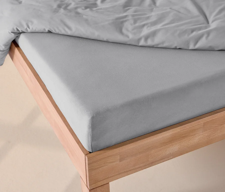 Un lit avec un drap-housse en jersey gris sur le matelas et une couette grise sur un cadre en bois clair.