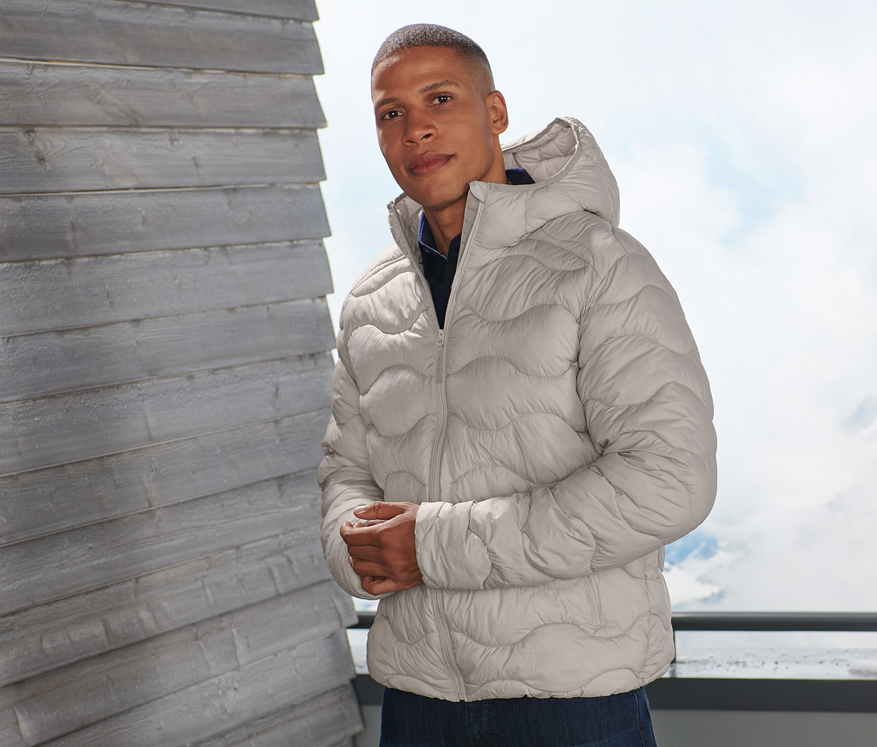 Un homme pose sur un balcon portant une veste matelassée beige.
