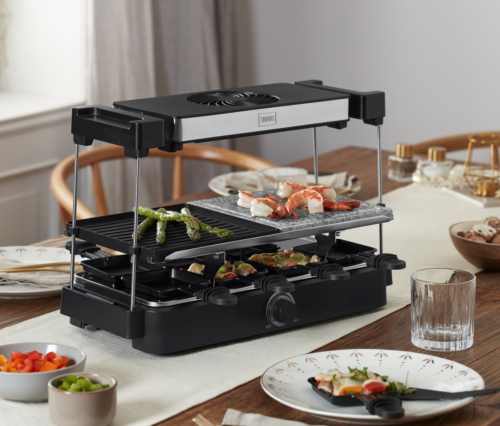 Sur une table en bois, un four à raclette Trebs « 15100 » avec hotte aspirante et plaque de pierre est en train de griller des aliments.