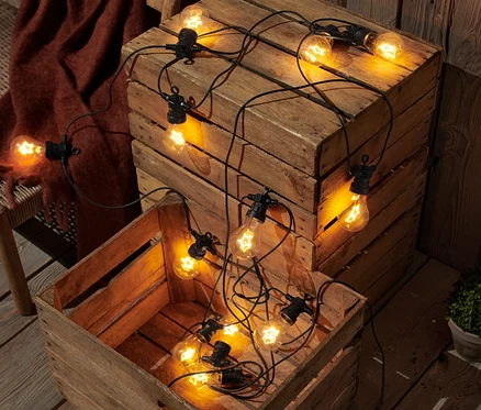 Deux caisses en bois avec des guirlandes lumineuses à filament LED sur une terrasse en bois à côté d'un banc en osier avec une couverture marron.
