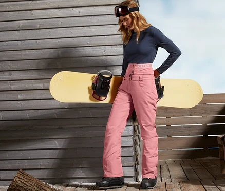Une femme se tient près d'un mur en bois et s'appuie sur un snowboard. Elle porte un pantalon de ski et de snowboard.