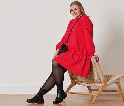 Une femme est assise sur une chaise en osier et porte une robe sweat rouge, des bottines en cuir, 2 paires de collants et des mini sacs.