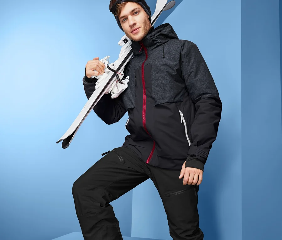 Un homme portant une veste de ski « High-End », un pantalon de ski, des chaussures de ski et des lunettes de ski porte des skis sur l'épaule.