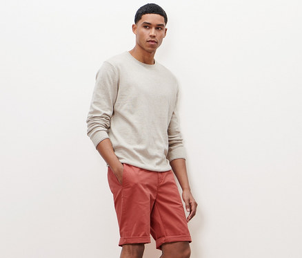 Un homme pose en pull beige et short chino terracotta.