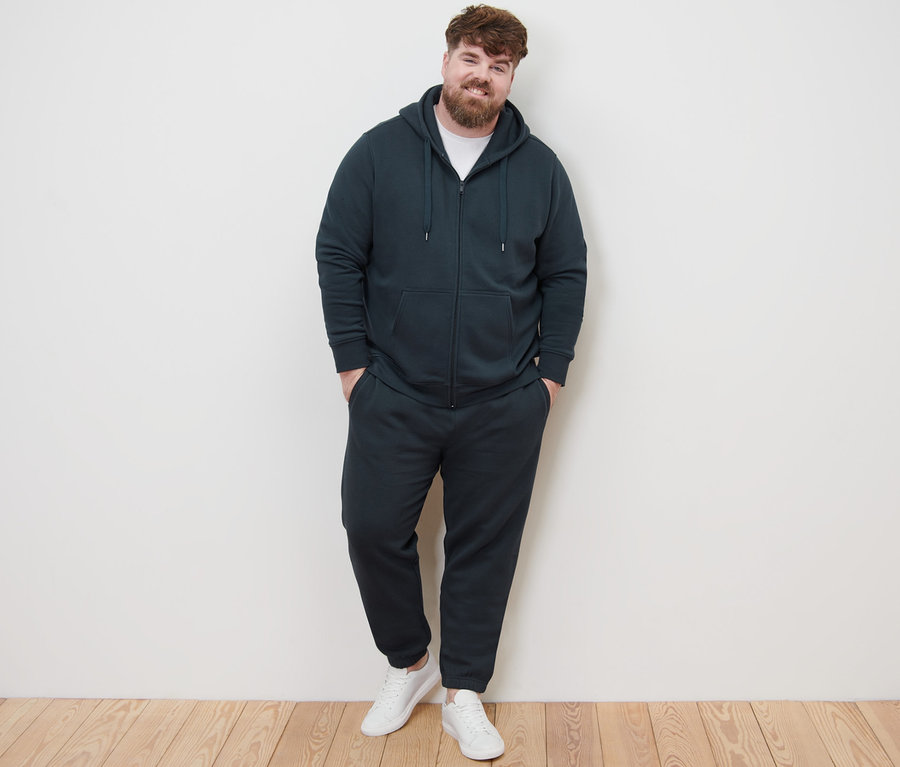 Un homme pose face à la caméra et porte une veste à capuche, un pantalon de survêtement et des baskets blanches.