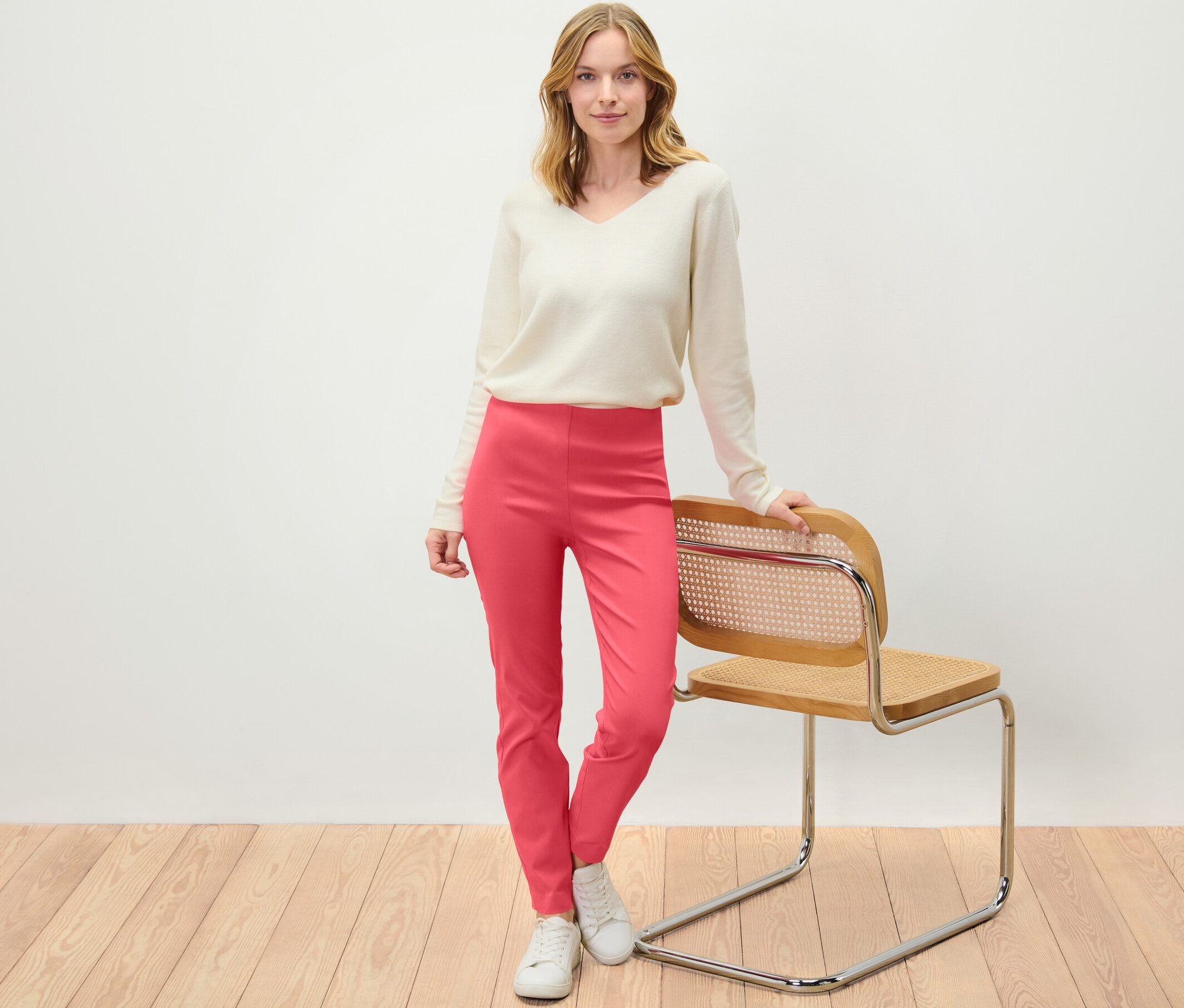 Une femme debout, appuyée sur une chaise, porte un pantalon stretch corail.