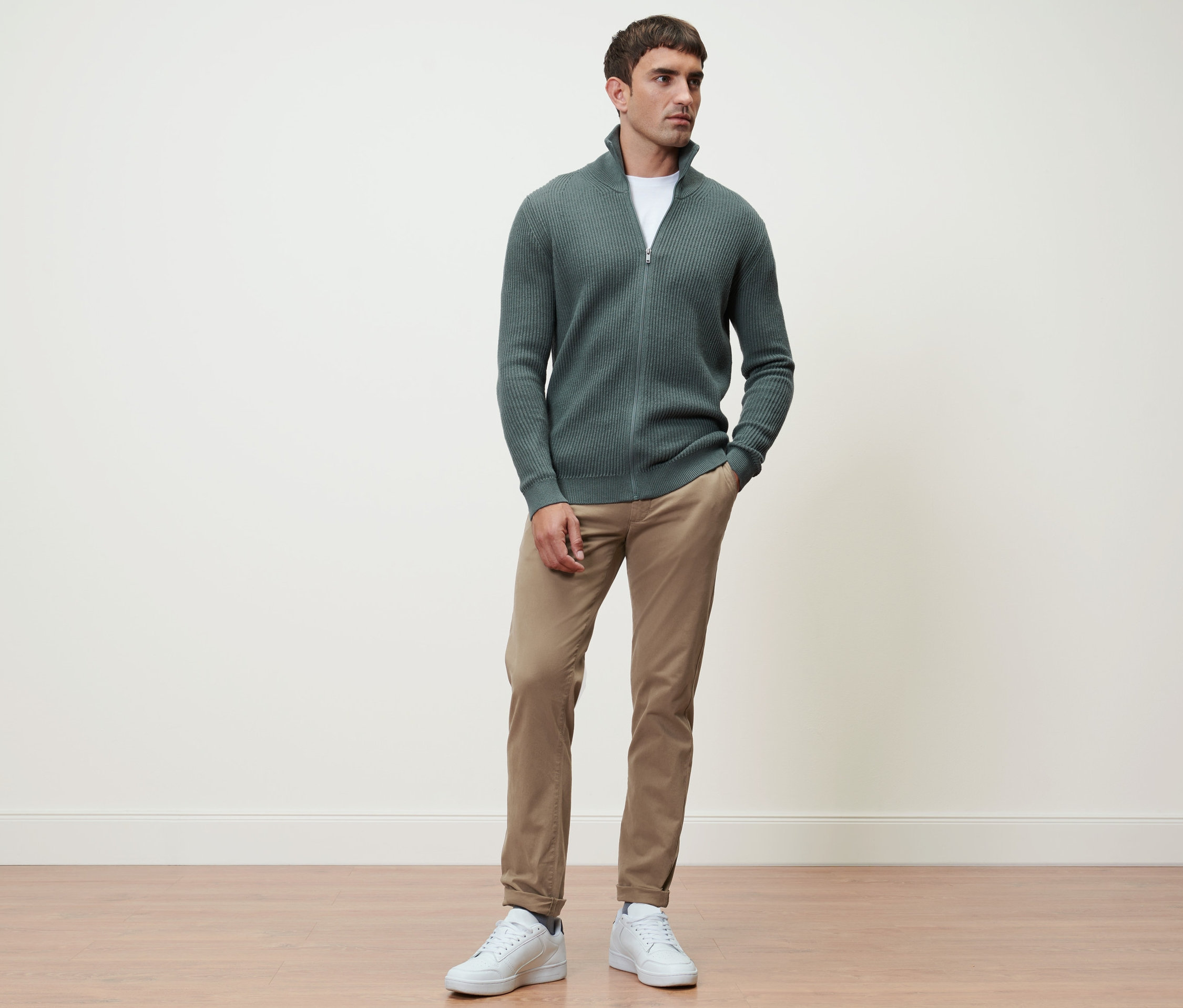 Un homme debout portant une jaquette en maille sauge et un pantalon chino sable.