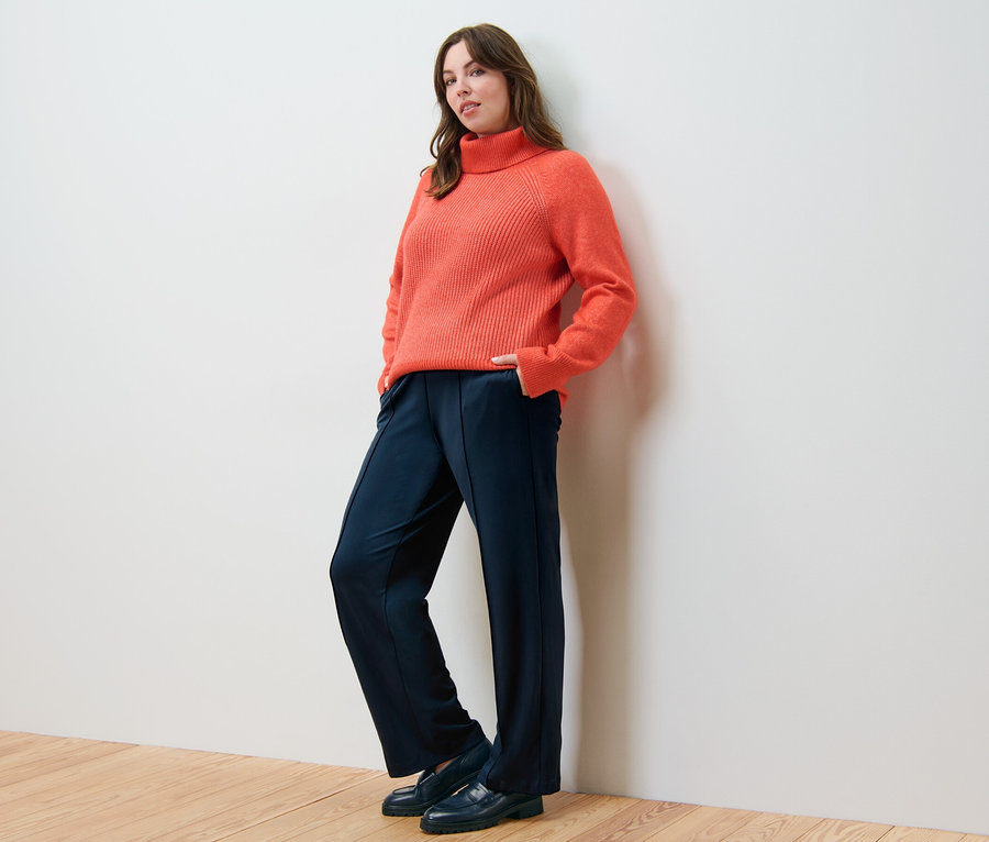 Femme posant en pull à col roulé orange en grosse maille et pantalon en jersey avec passepoil.