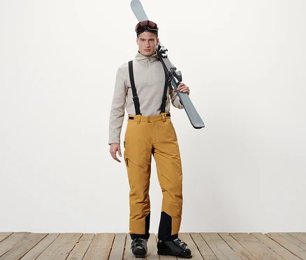 Un homme debout portant des skis sur l'épaule, un pull beige et un pantalon de ski camel.