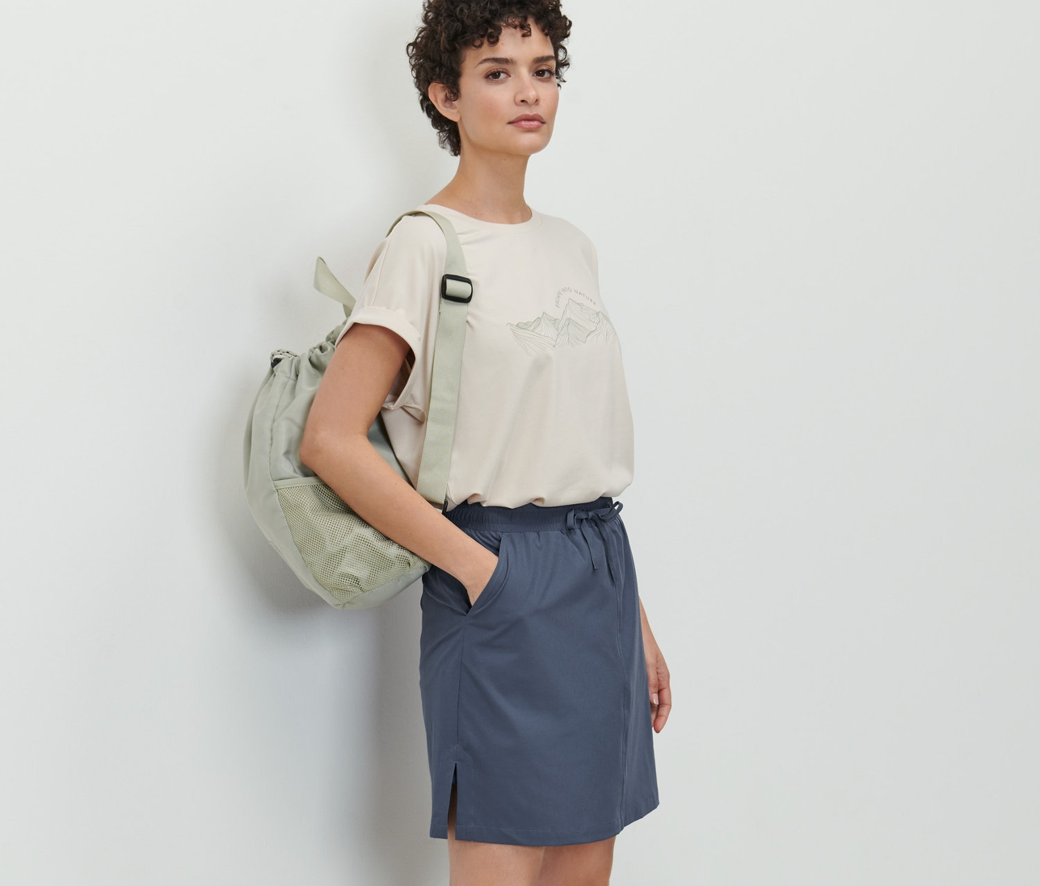 Une femme pose, portant une jupe fonctionnelle bleu foncé avec un short intégré, associée à un t-shirt couleur crème et un sac à dos vert.