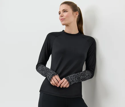 Une femme appuyée contre un mur porte un t-shirt technique à manches longues noir et argenté.