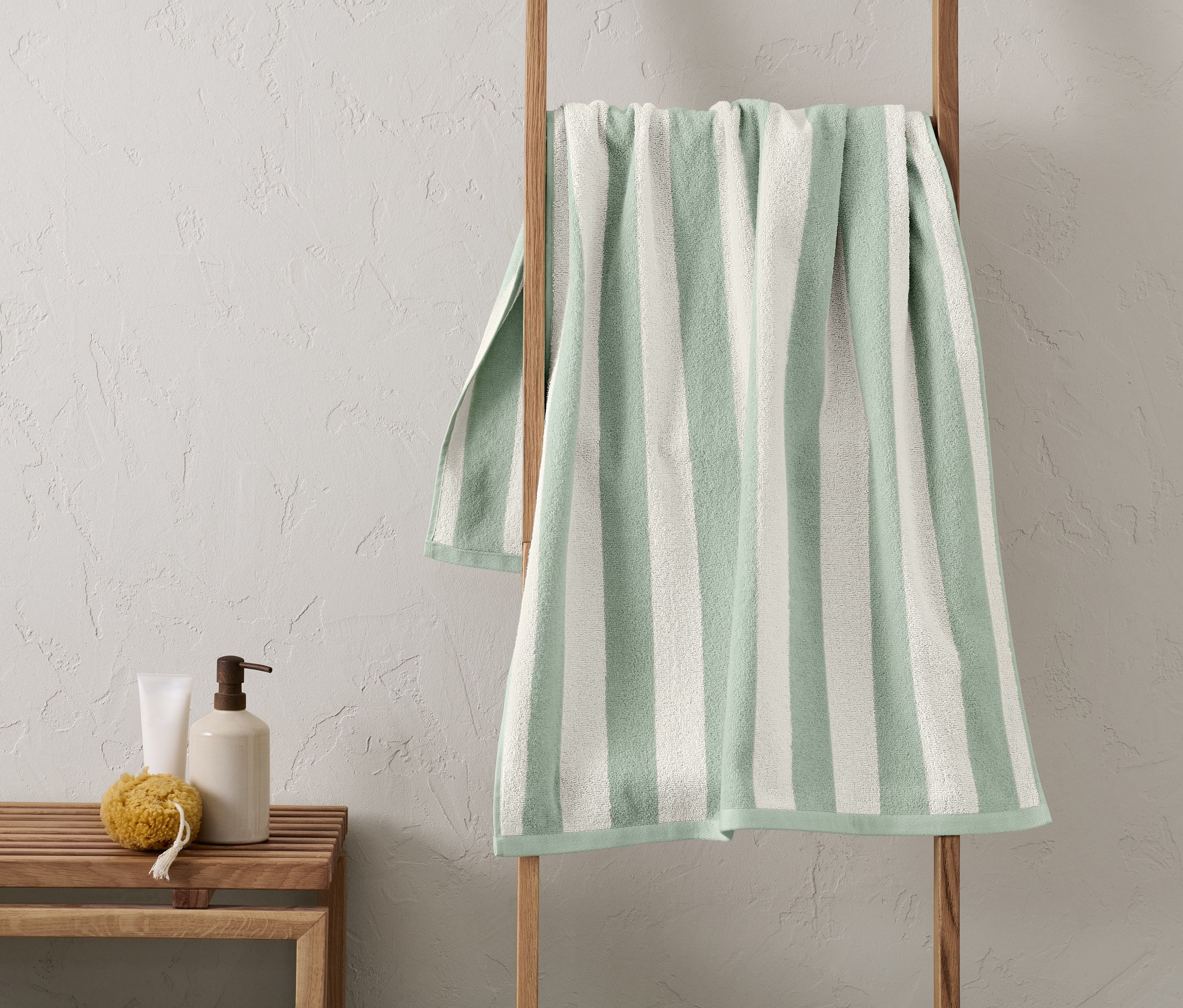 Un drap de bain rayé vert est suspendu à une échelle en bois. À côté de l'échelle, il y a un banc en bois avec une bouteille et une éponge.