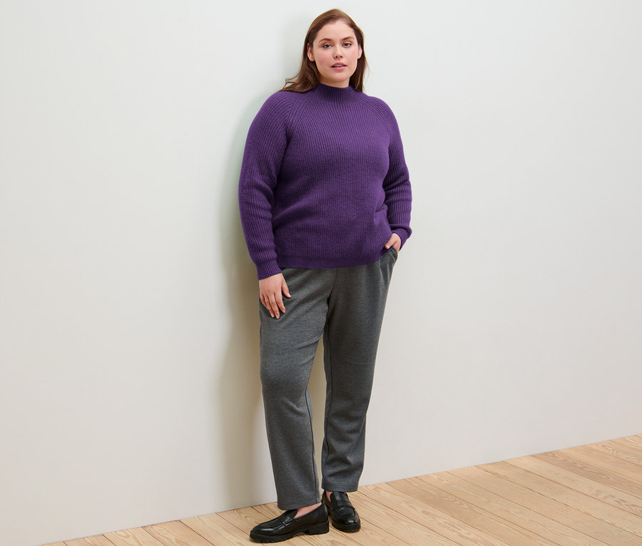 Une femme debout, les mains dans les poches de son pantalon de jogging, porte un pull violet à col droit en grosse maille.