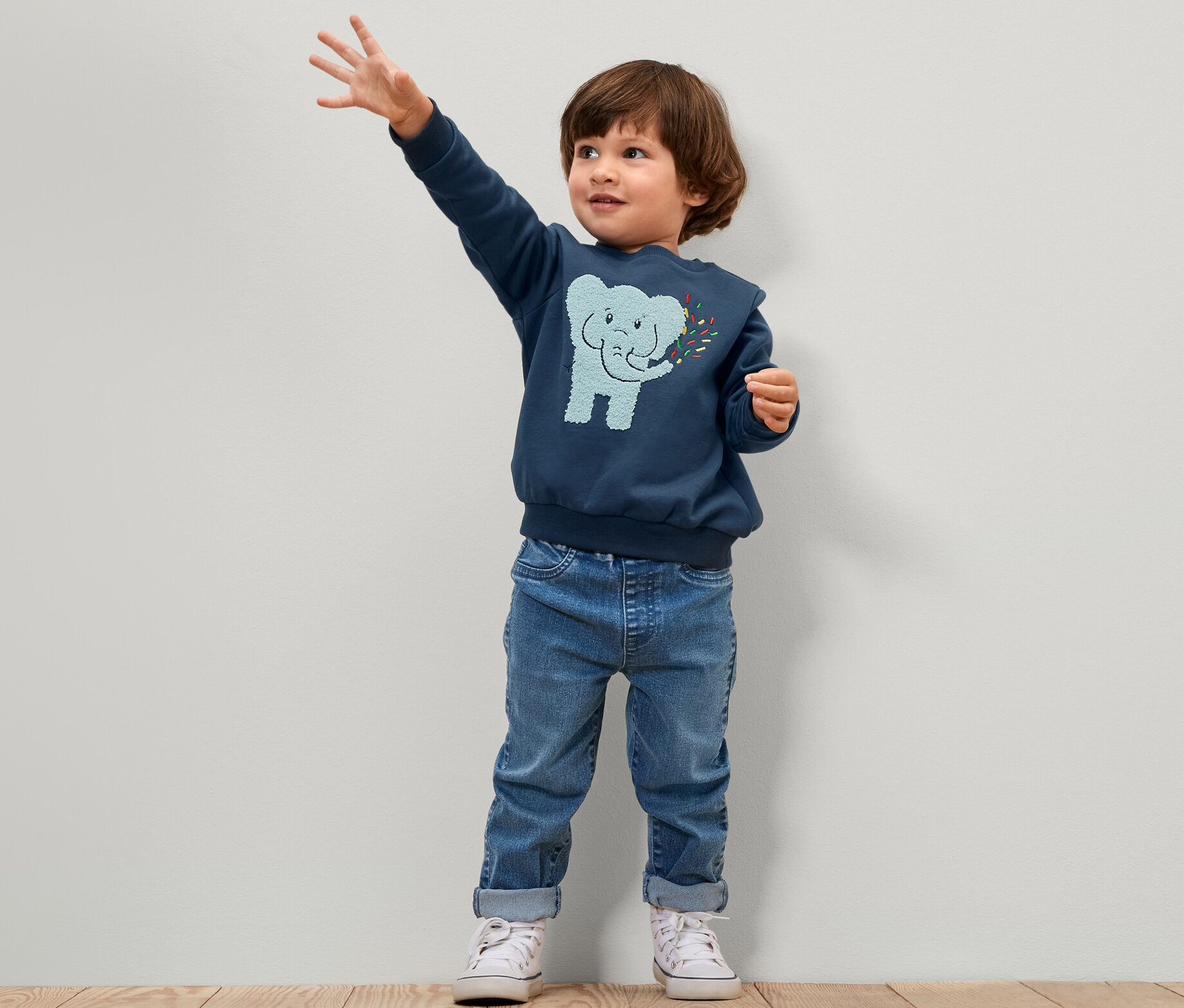 Un garçon se tient debout et fait un signe de la main. Il porte un pantalon à enfiler bleu pour enfant – Fit « Luca » et un pull bleu avec un éléphant.